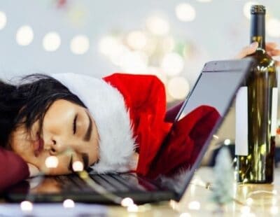 Image: Woman in Santa hat asleep on her laptop holding a bottle of wine. How can I drink less this Christmas?