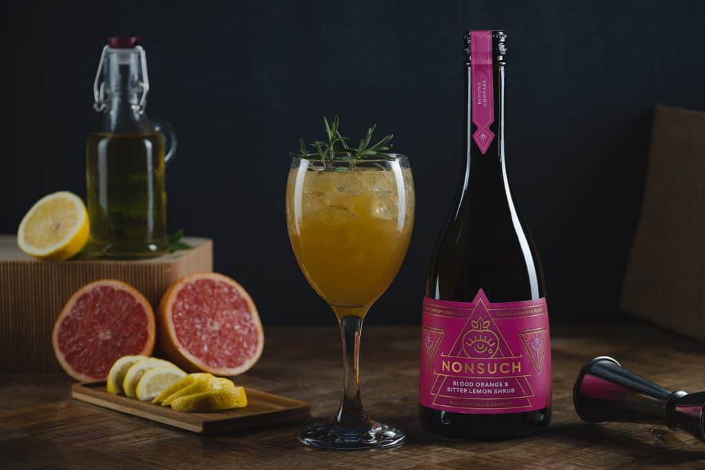 A non-alcoholic cocktail in a wine glass, garnished with rosemary with a blood orange cut open next to it and a bottle of Nonsuch Shrubs